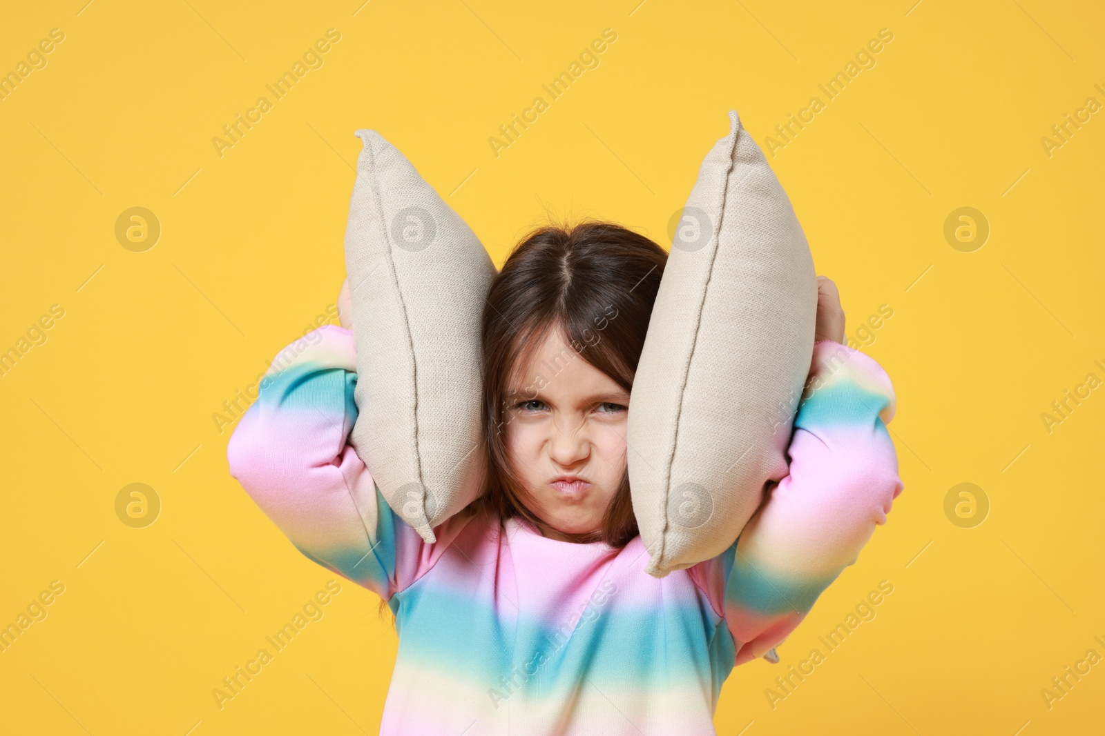 Photo of Annoyed little girl covering her ears with pillows from loud sound on yellow background