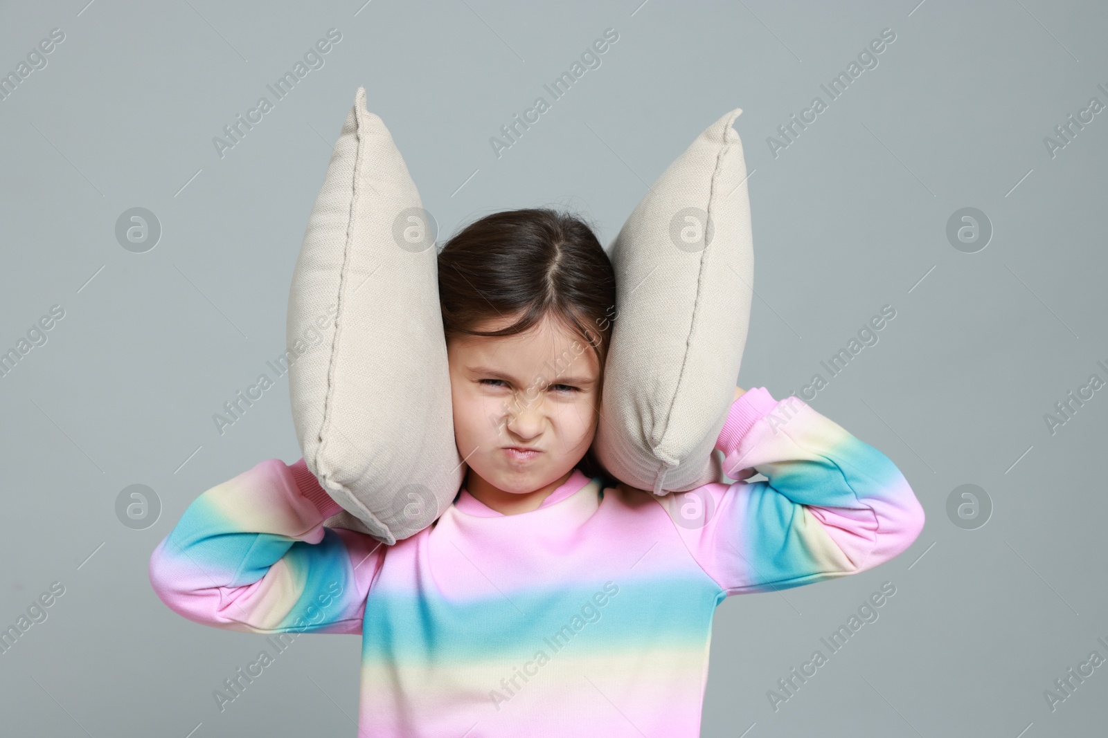 Photo of Annoyed little girl covering her ears with pillows from loud sound on grey background