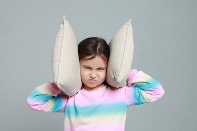 Photo of Annoyed little girl covering her ears with pillows from loud sound on grey background