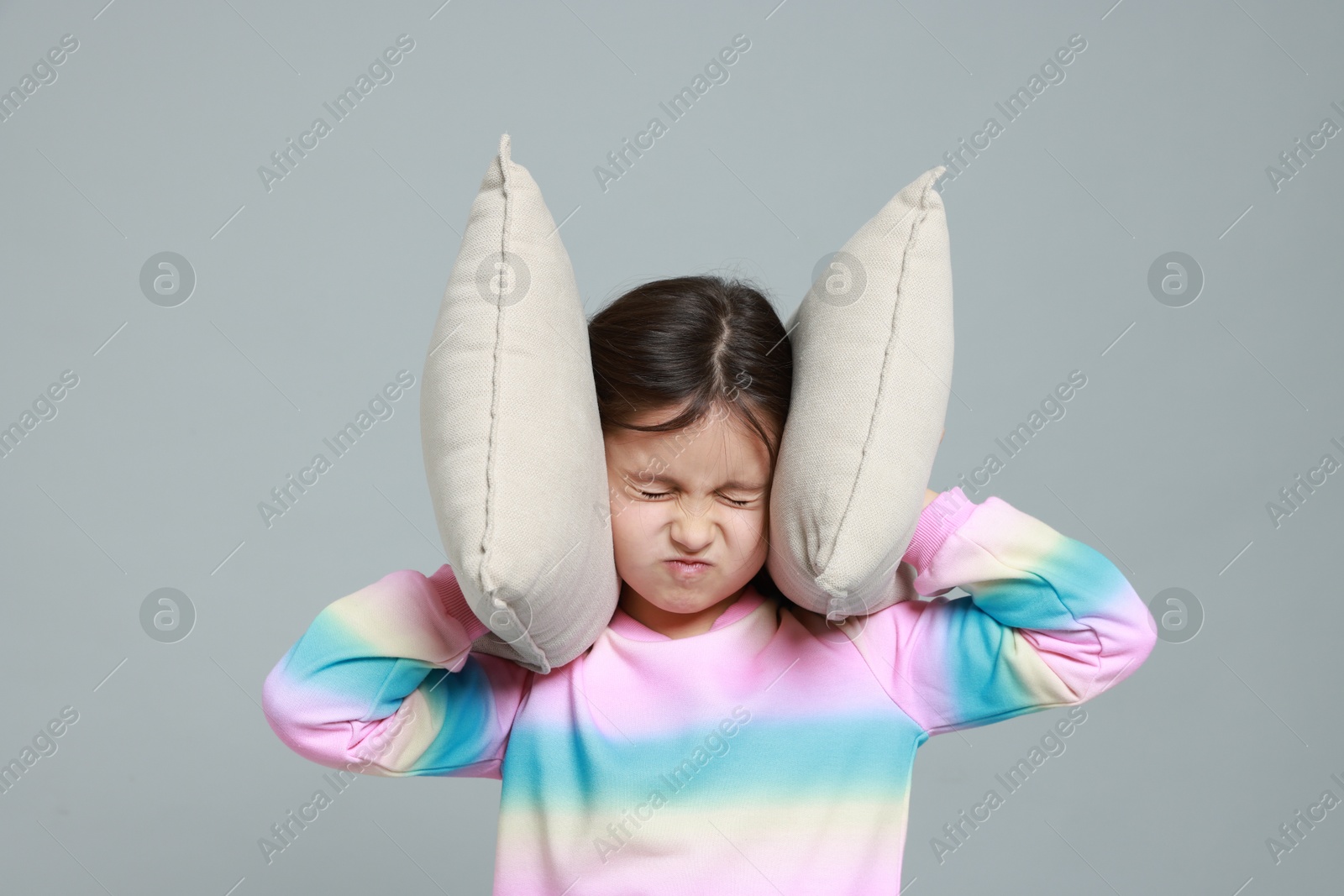 Photo of Annoyed little girl covering her ears with pillows from loud sound on grey background