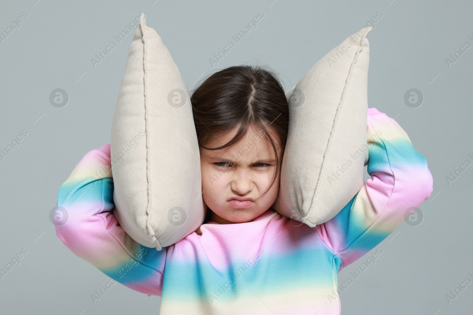 Photo of Annoyed little girl covering her ears with pillows from loud sound on grey background