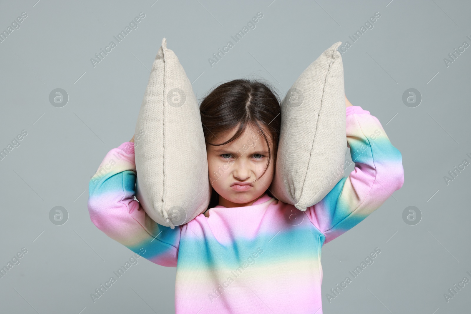 Photo of Annoyed little girl covering her ears with pillows from loud sound on grey background