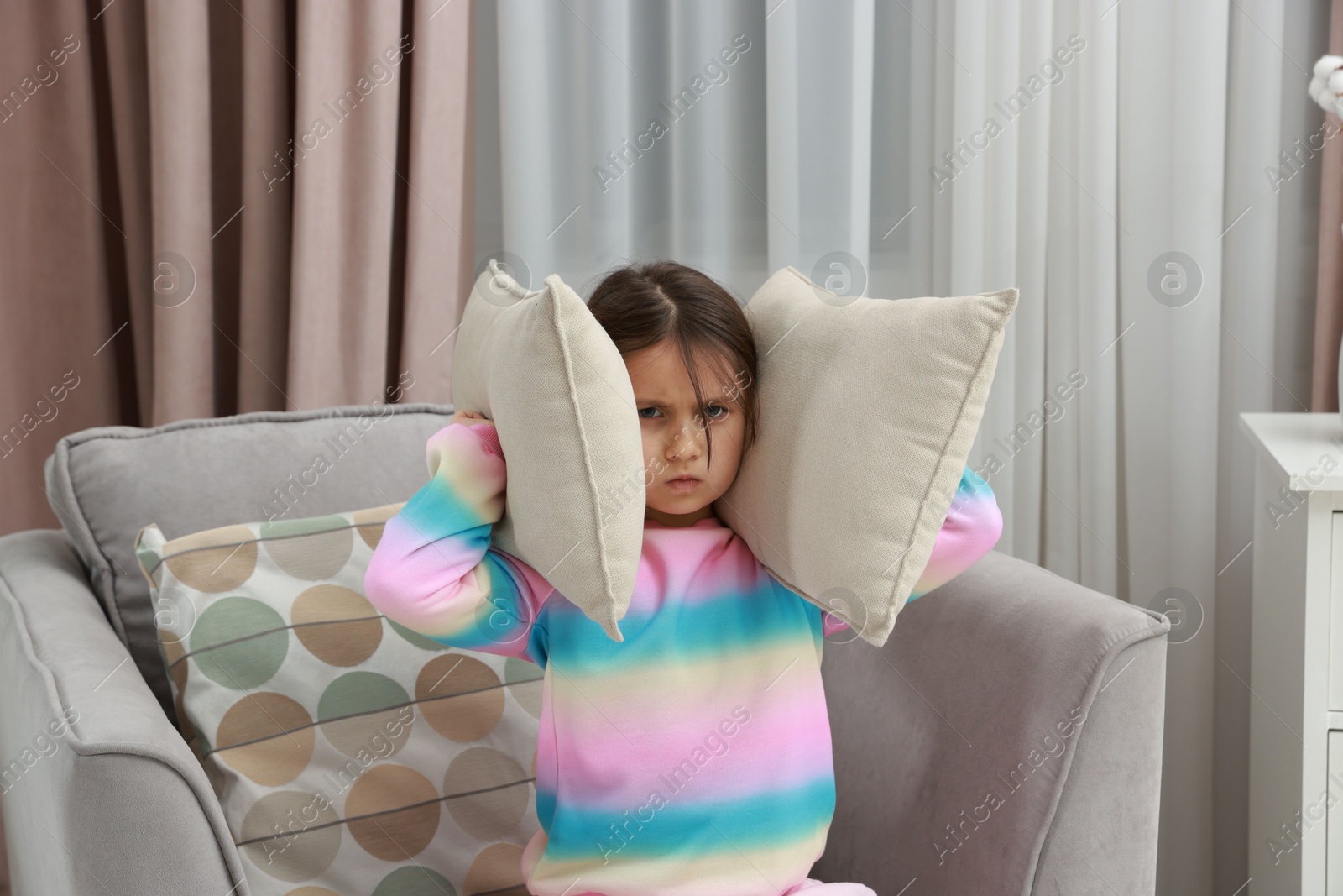 Photo of Angry little girl covering her ears with pillows from loud sound on armchair at home