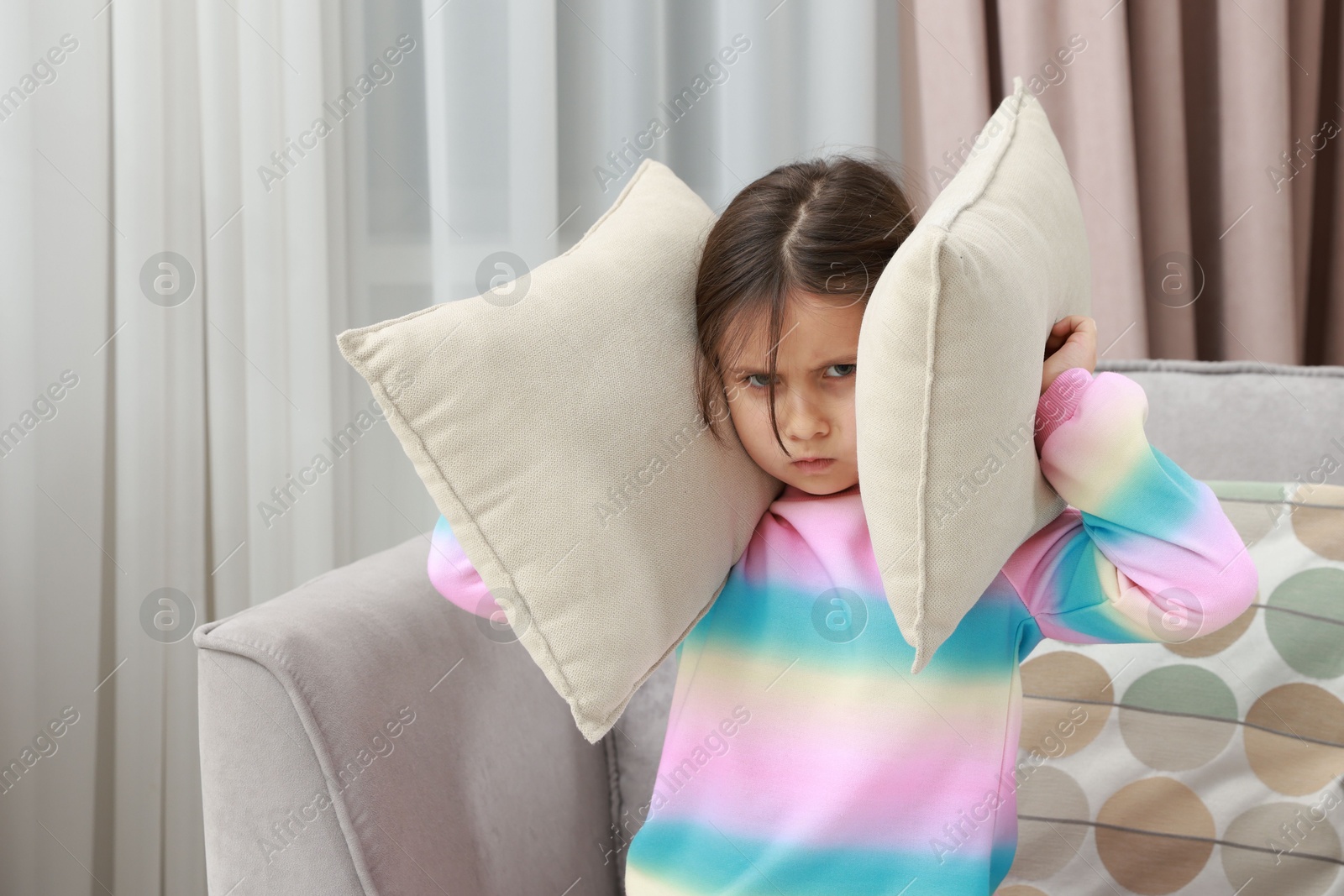 Photo of Angry little girl covering her ears with pillows from loud sound on armchair at home