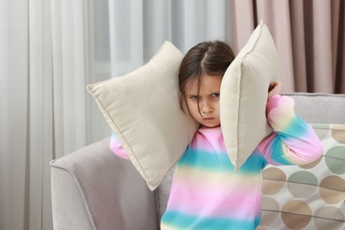 Photo of Angry little girl covering her ears with pillows from loud sound on armchair at home