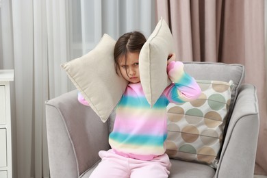 Photo of Angry little girl covering her ears with pillows from loud sound on armchair at home