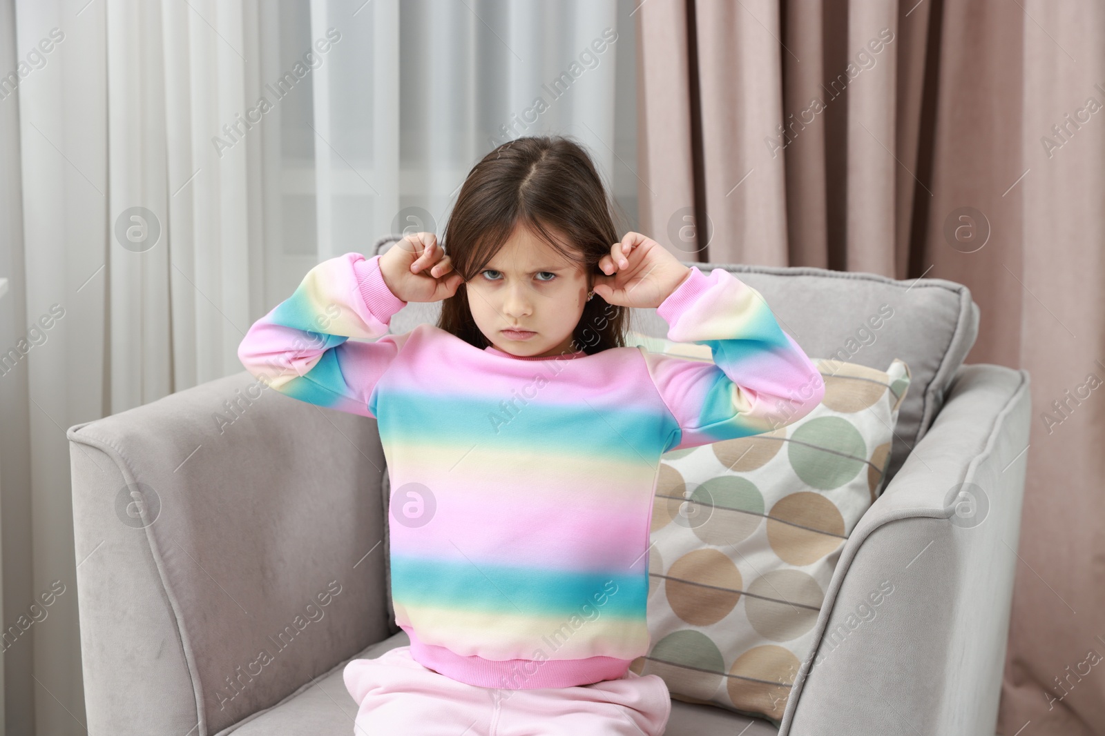 Photo of Angry little girl covering her ears from loud sound on armchair at home