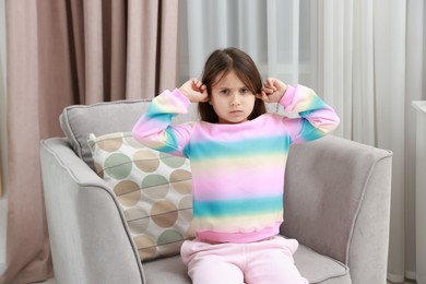 Photo of Angry little girl covering her ears from loud sound on armchair at home