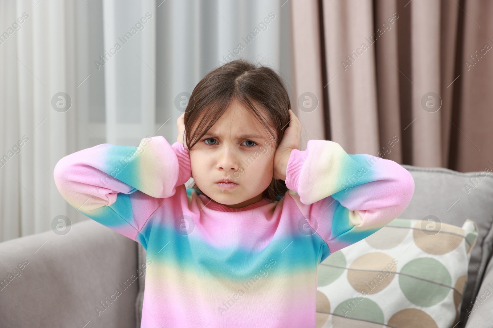 Photo of Annoyed little girl covering her ears with pillows from loud sound on armchair at home