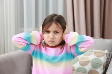 Photo of Annoyed little girl covering her ears with pillows from loud sound on armchair at home