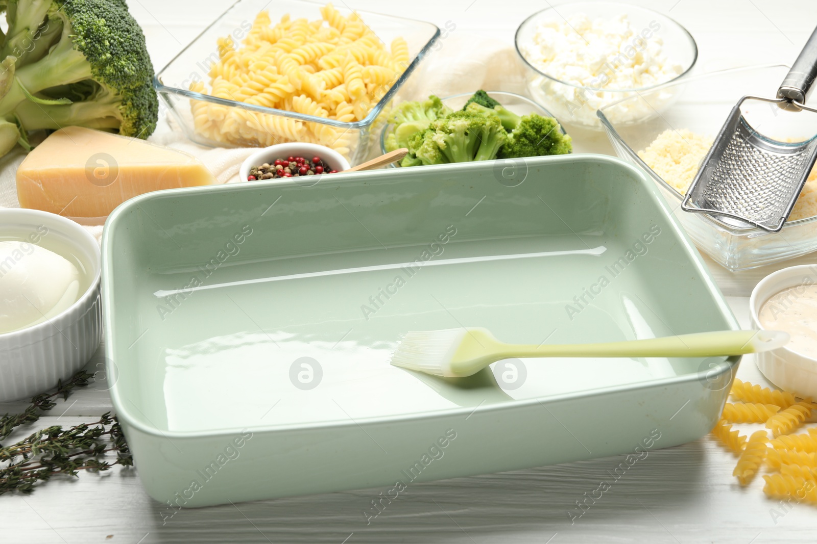 Photo of Ceramic casserole, brush and ingredients on white wooden table, closeup