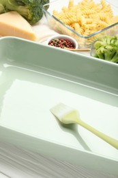 Photo of Ceramic casserole, brush and ingredients on white wooden table, closeup