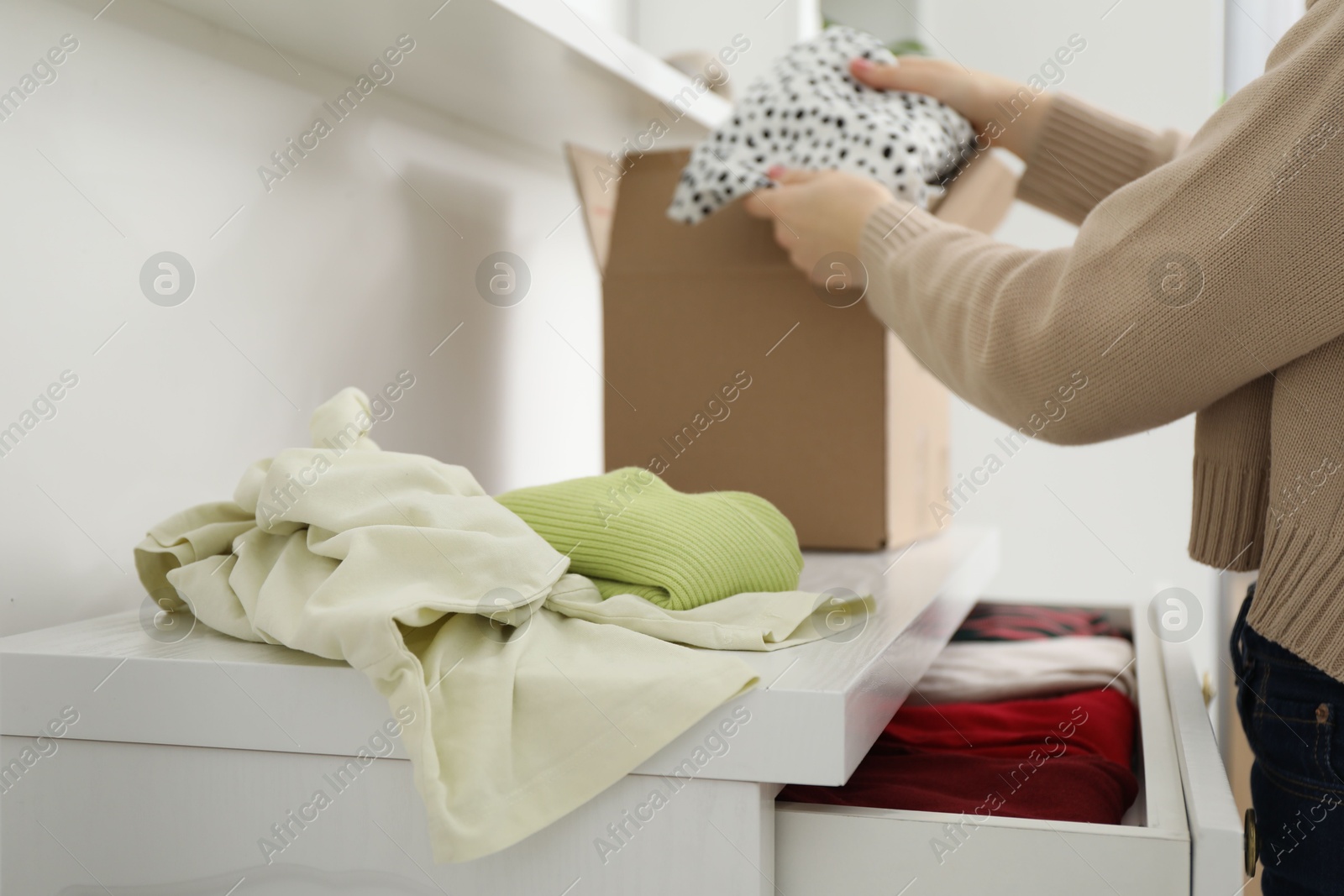 Photo of Woman stacking used clothes into box at chest of drawers indoors, closeup