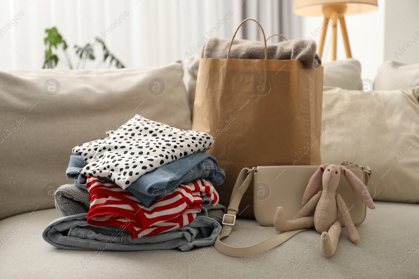 Photo of Stack of different used clothes, toy and paper bag on sofa indoors