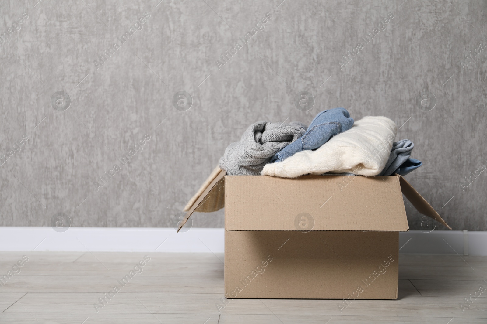 Photo of Used clothes in cardboard box on floor near grey wall. Space for text