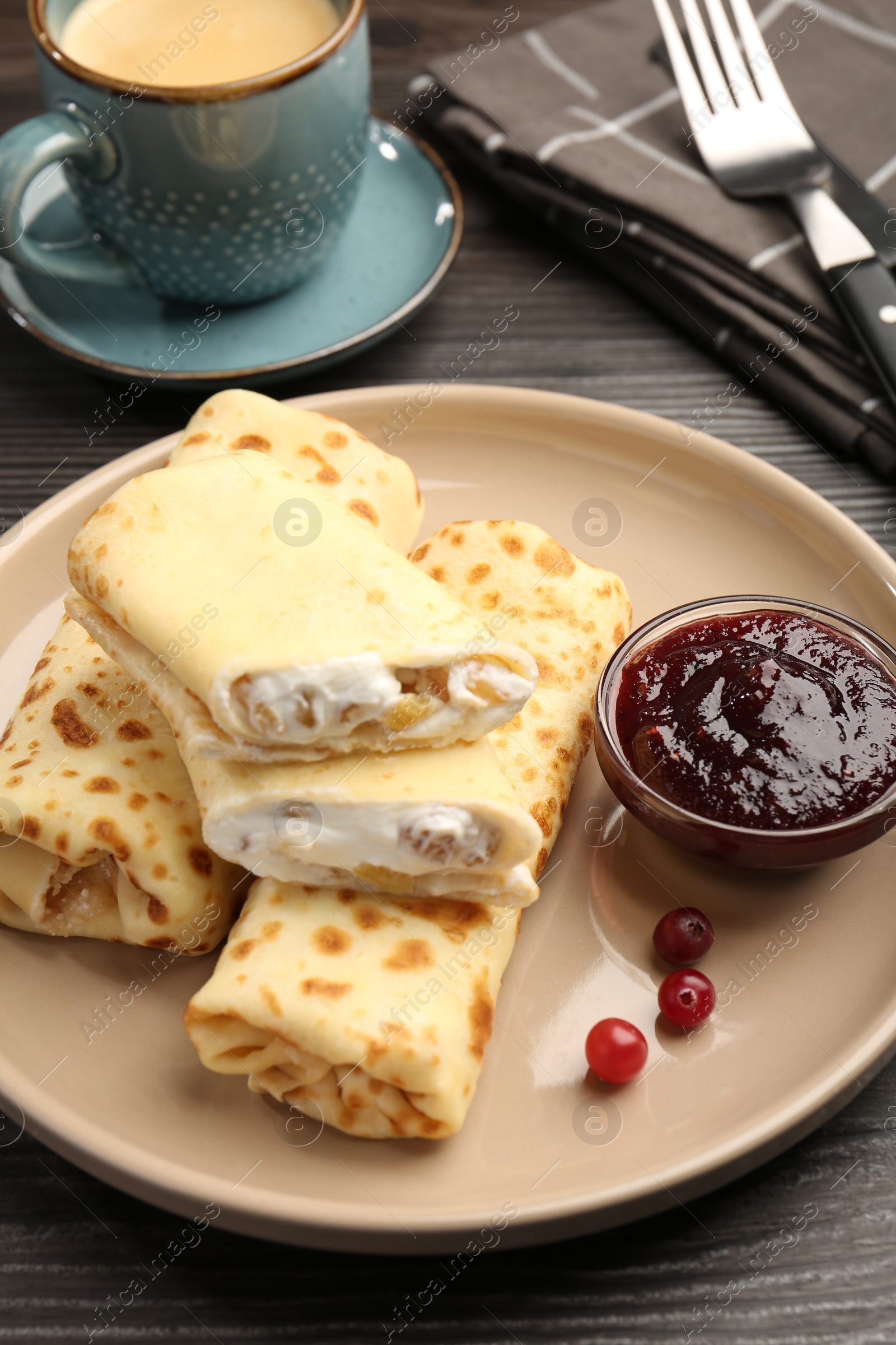 Photo of Delicious rolled crepes with cottage cheese, raisins, jam, cranberries and coffee on wooden table, closeup