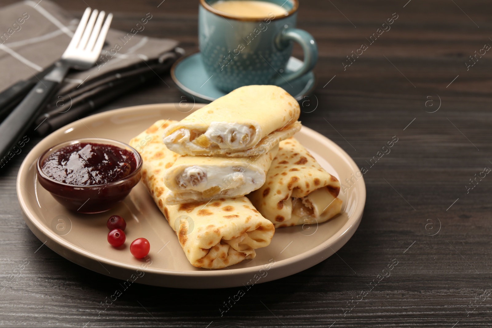 Photo of Delicious rolled crepes with cottage cheese, raisins, jam, cranberries and coffee on wooden table, closeup