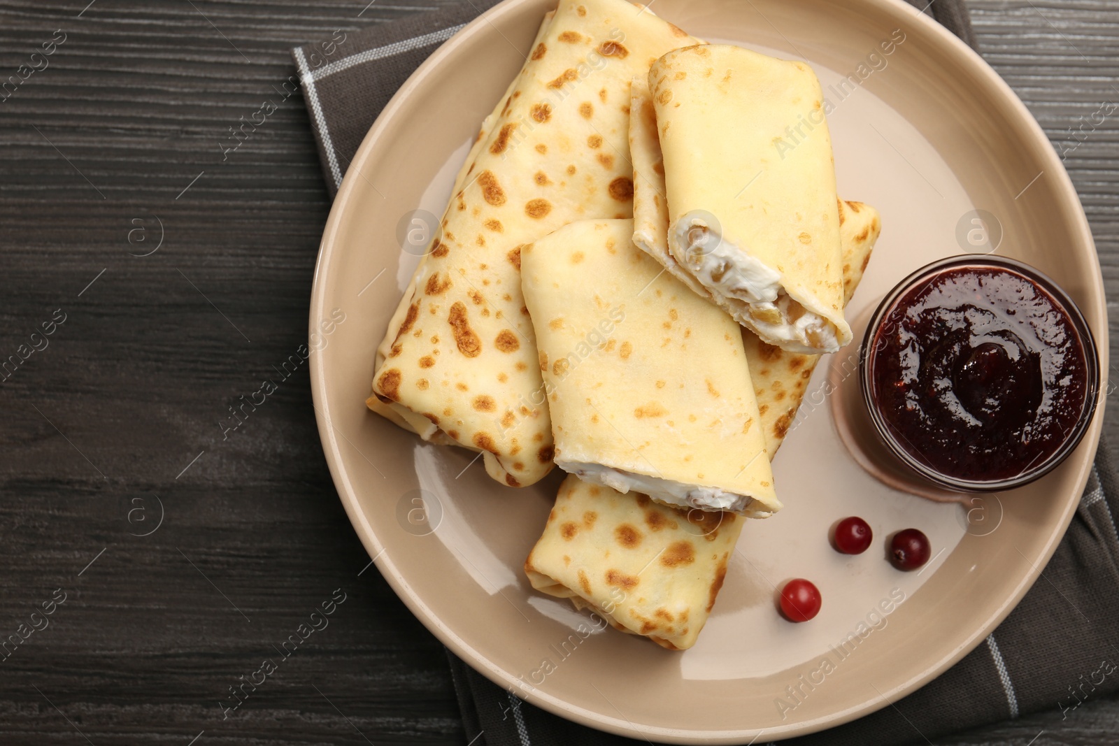 Photo of Delicious rolled crepes with cottage cheese, raisins, jam and cranberries on wooden table, top view