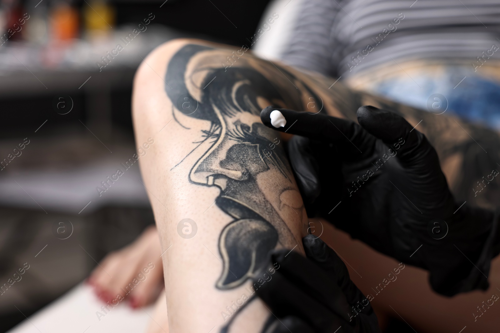 Photo of Professional artist in gloves applying cream onto woman's leg with fresh tattoo in salon, closeup