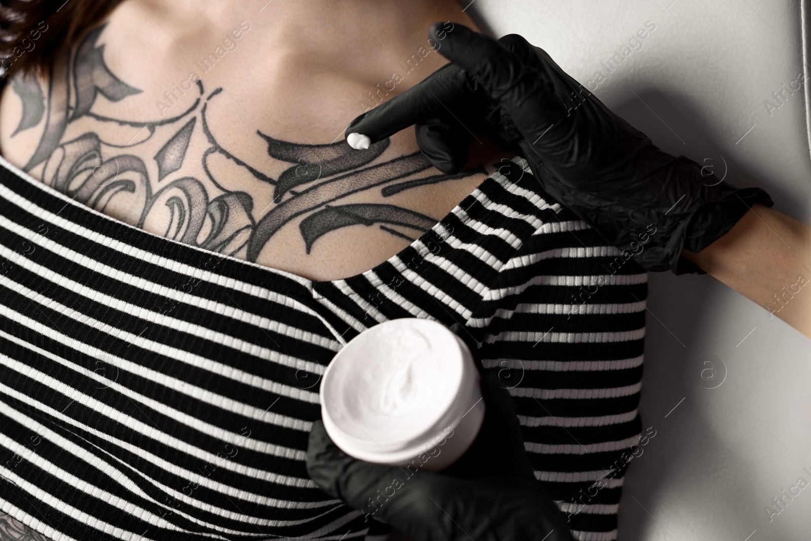 Photo of Professional artist in gloves applying cream onto woman's skin with fresh tattoo in salon, closeup