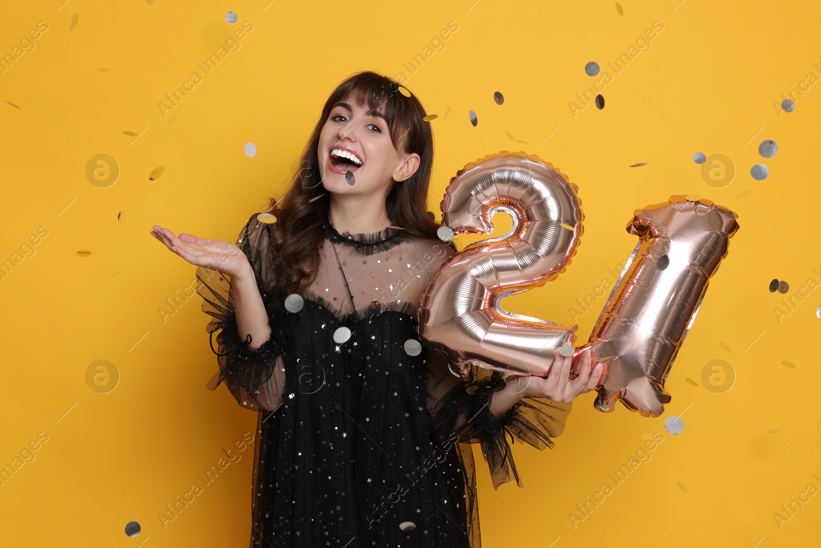 Photo of Coming of age party - 21st birthday. Young woman holding number shaped balloons on yellow background with confetti