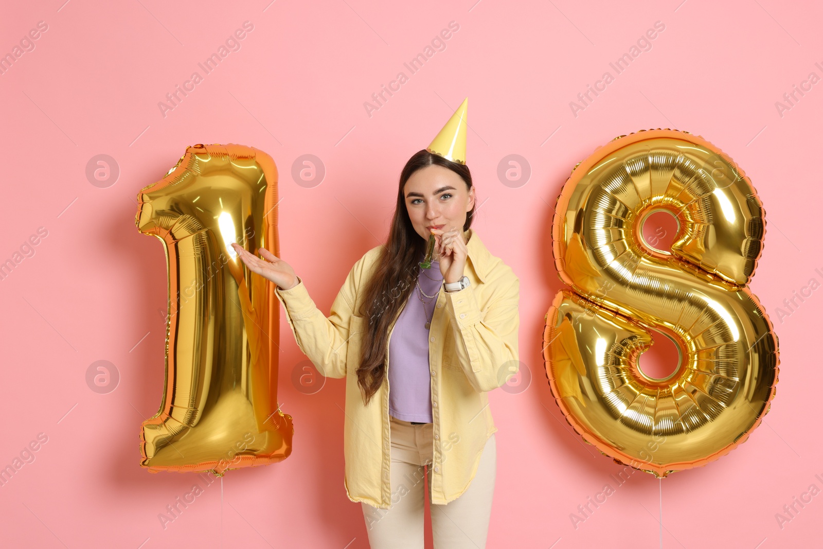 Photo of Coming of age party - 18th birthday. Happy young woman with number shaped balloons, hat and blower on pink background