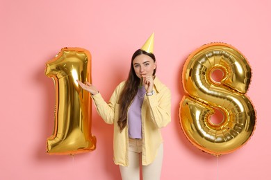 Photo of Coming of age party - 18th birthday. Happy young woman with number shaped balloons, hat and blower on pink background