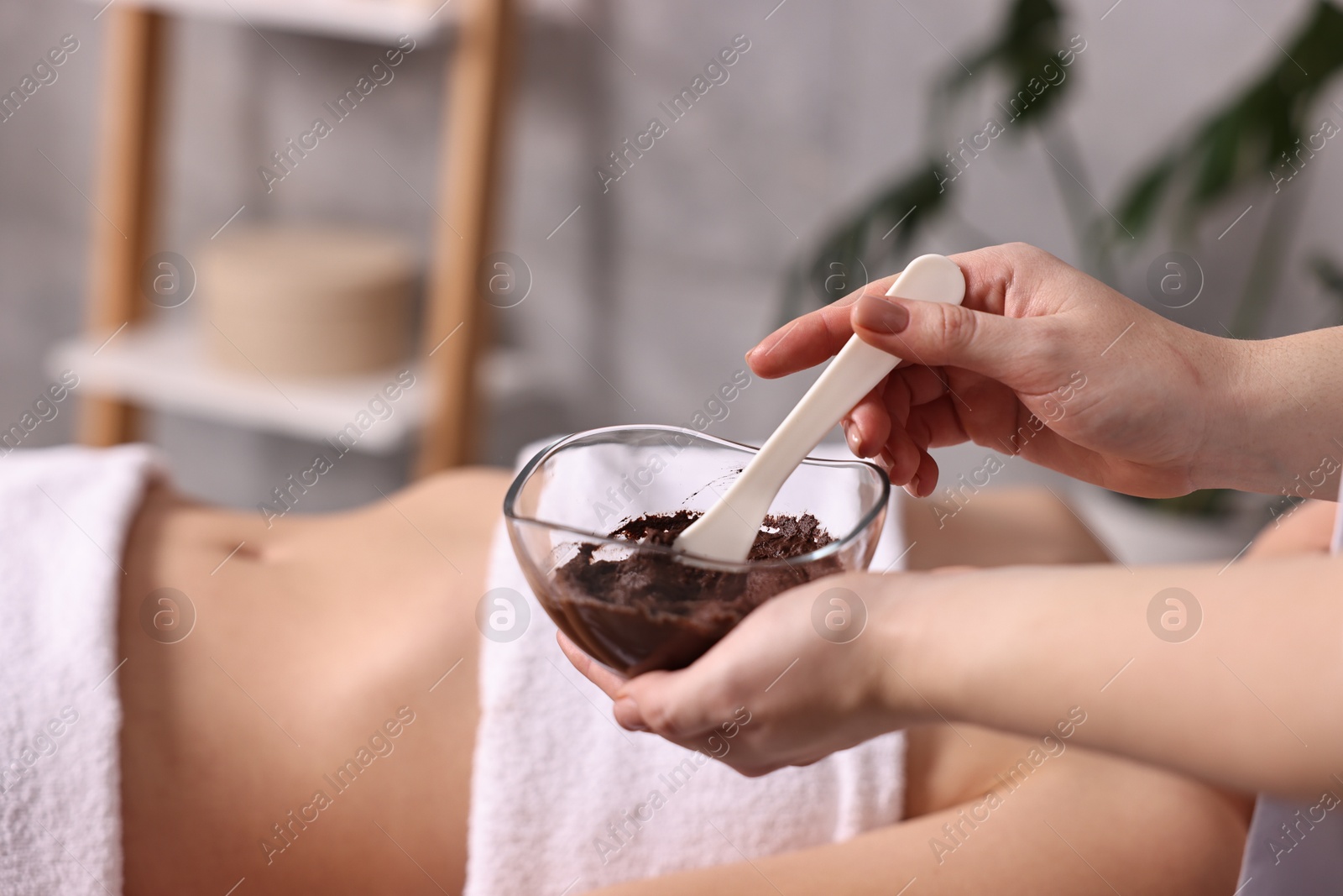 Photo of Spa worker with cosmetic product for chocolate body wrap near woman in salon, closeup