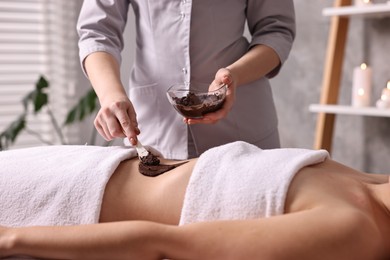 Photo of Chocolate body wrap. Spa worker applying mask onto woman's belly in salon, closeup