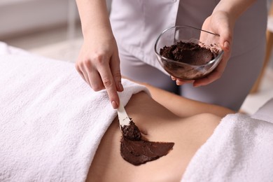 Photo of Chocolate body wrap. Spa worker applying mask onto woman's belly in salon, closeup