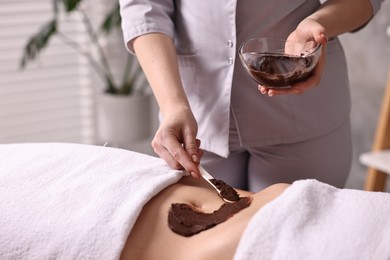 Photo of Chocolate body wrap. Spa worker applying mask onto woman's belly in salon, closeup