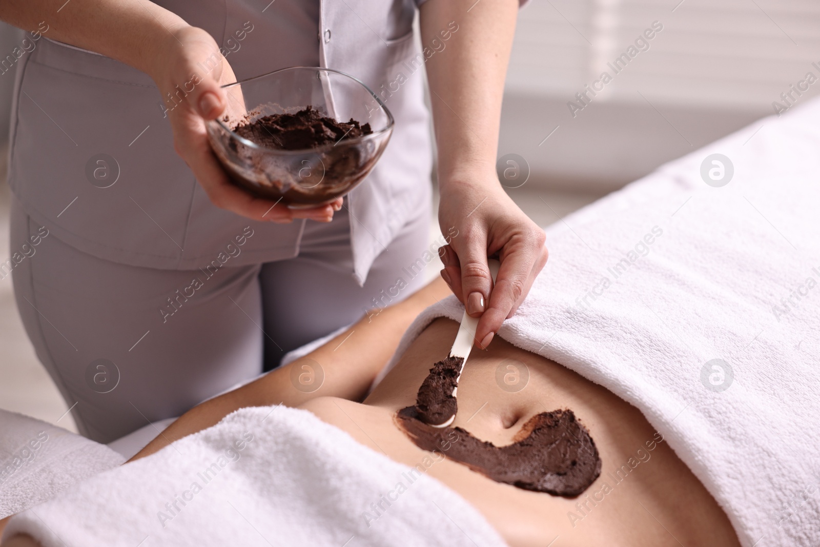 Photo of Chocolate body wrap. Spa worker applying mask onto woman's belly in salon, closeup