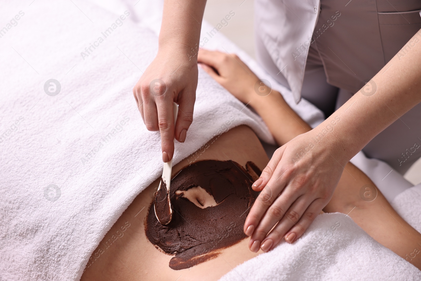 Photo of Chocolate body wrap. Spa worker applying mask onto woman's belly in salon, closeup