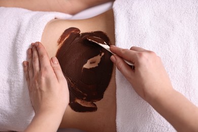 Photo of Chocolate body wrap. Spa worker applying mask onto woman's belly in salon, top view