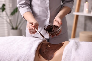 Photo of Chocolate body wrap. Spa worker applying mask onto woman's belly in salon, closeup