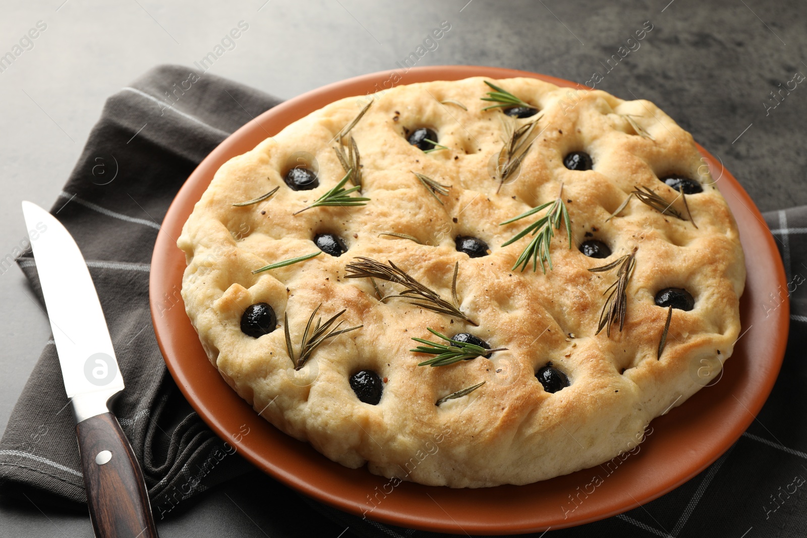 Photo of Delicious focaccia bread with olives, rosemary and knife on grey table, closeup