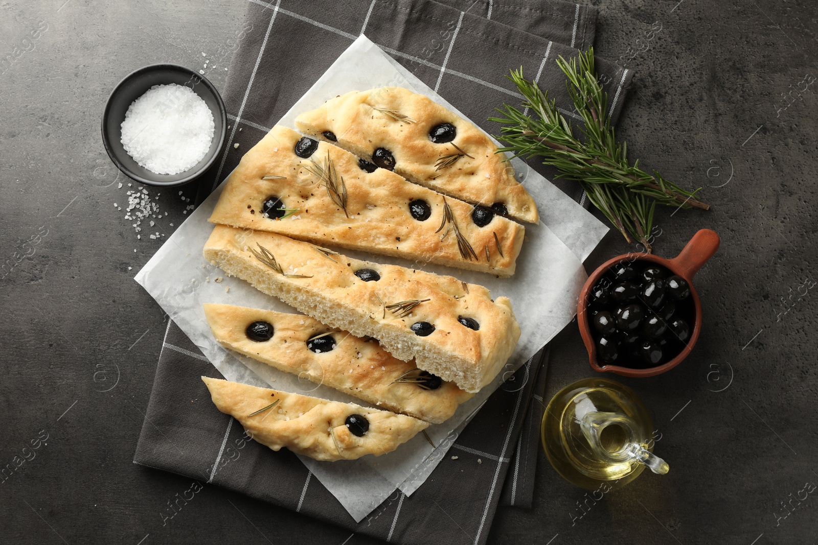 Photo of Slices of delicious focaccia bread with olives, rosemary, oil and salt on grey table, flat lay