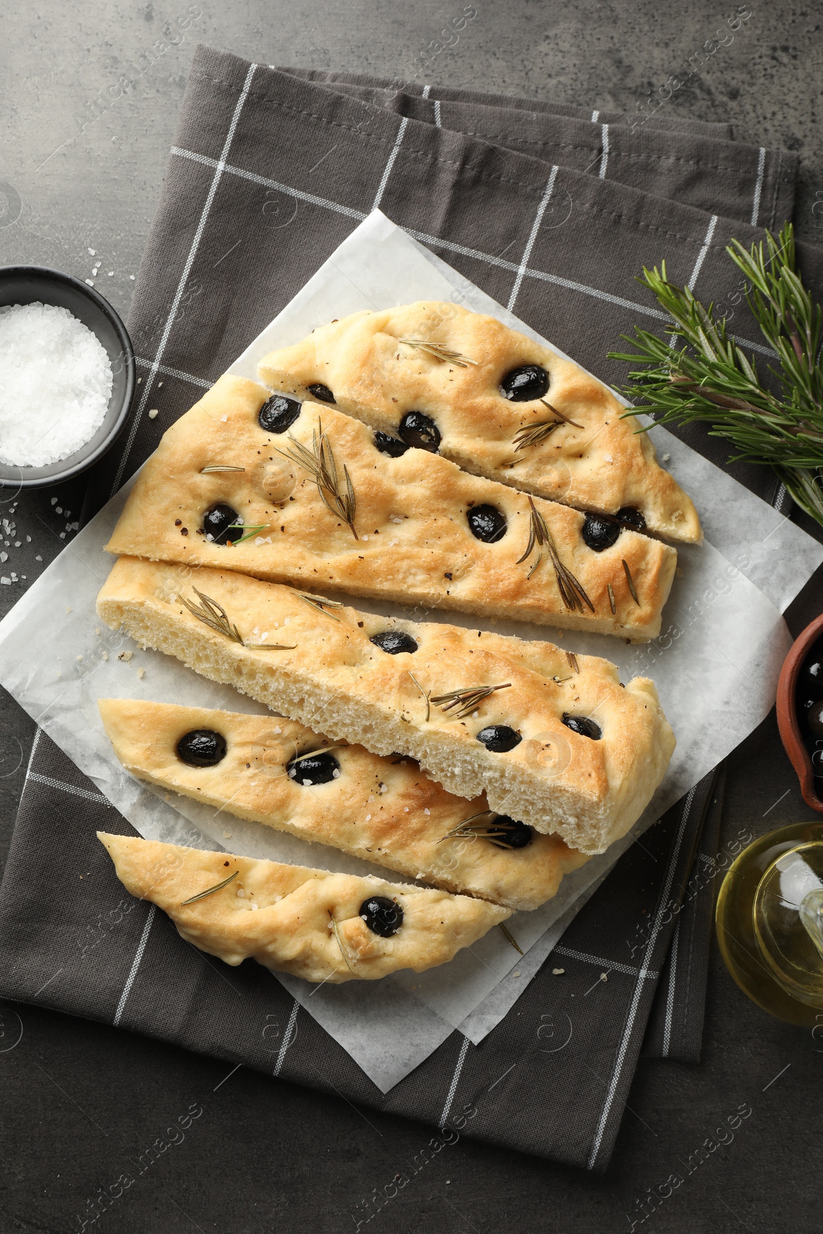 Photo of Slices of delicious focaccia bread with olives, rosemary, oil and salt on grey table, flat lay