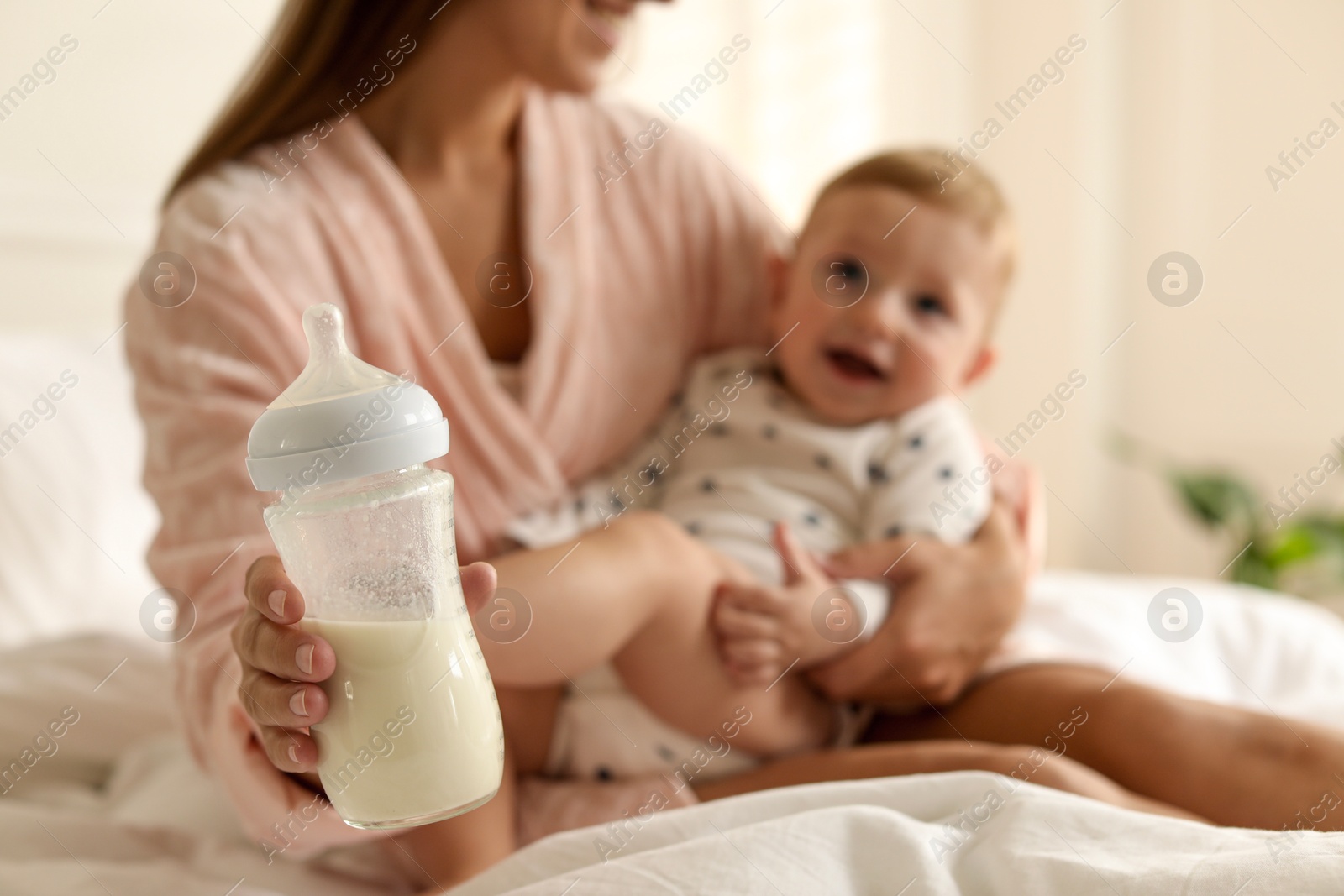 Photo of Mother holding cute little baby and bottle of milk on bed at home, selective focus