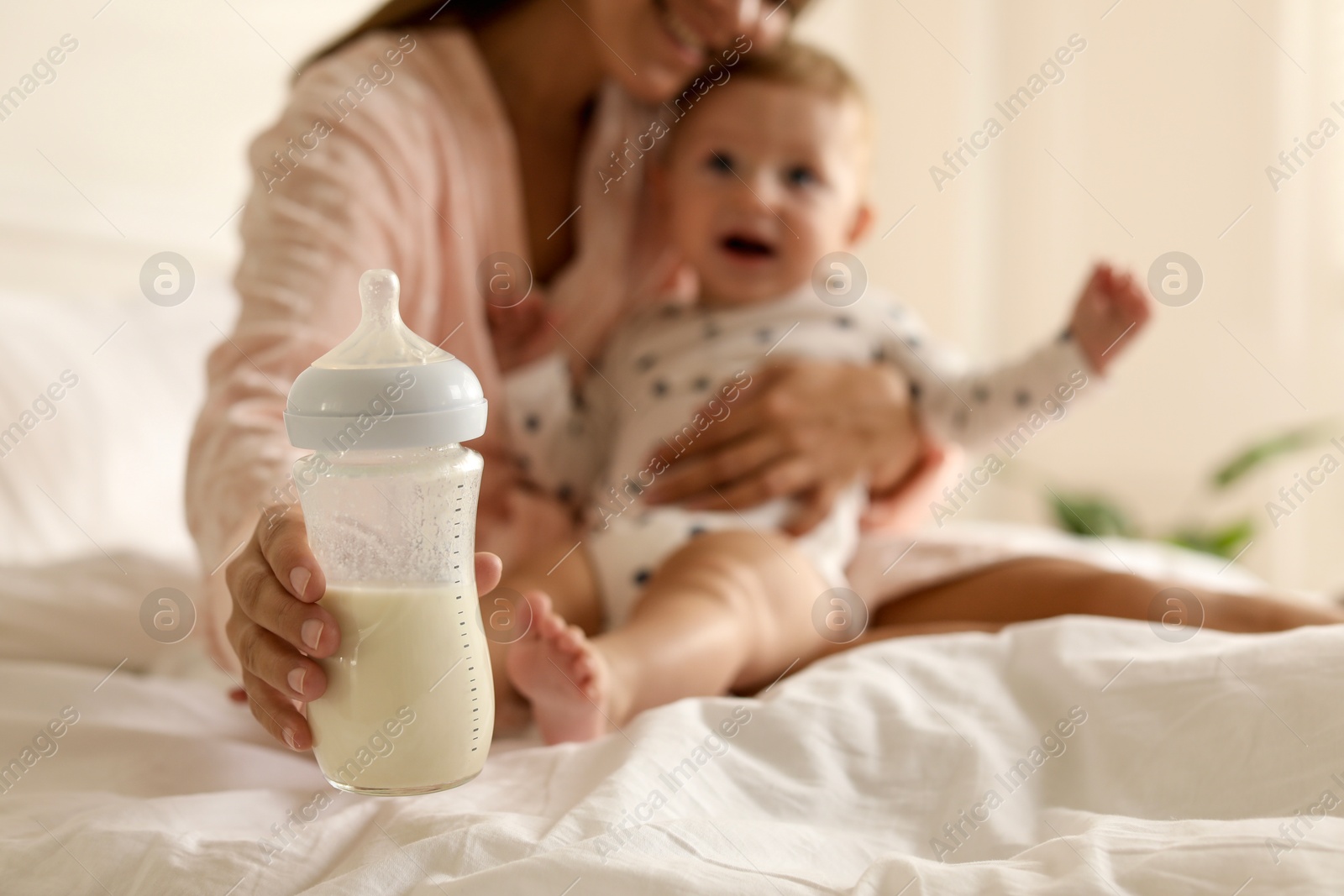 Photo of Mother holding cute little baby and bottle of milk on bed at home, selective focus