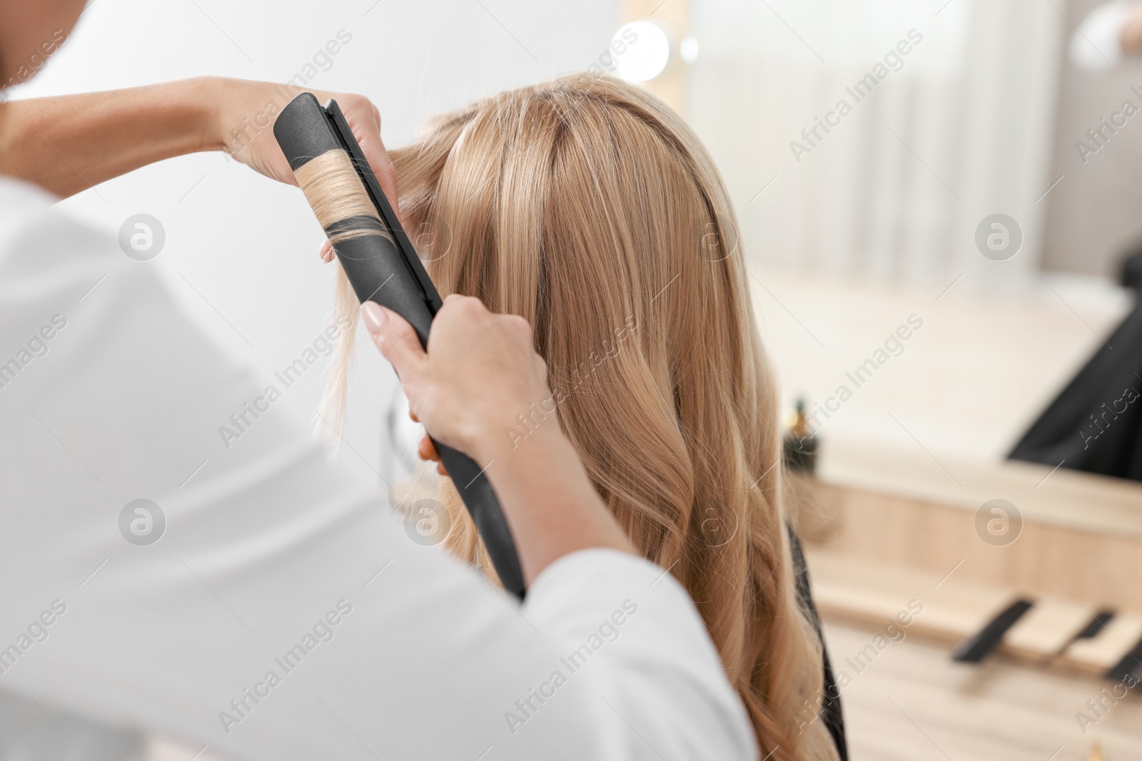 Photo of Hairdresser curling woman's hair with flat iron in salon, closeup