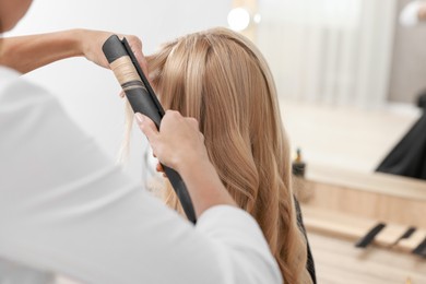 Photo of Hairdresser curling woman's hair with flat iron in salon, closeup