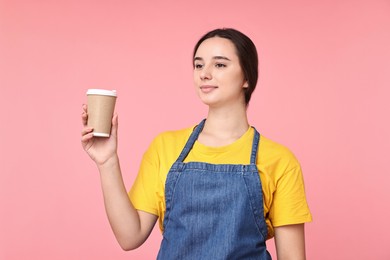 Photo of Girl in apron with paper cup of coffee on pink background. Work for teenagers