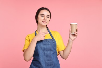 Photo of Girl in apron with paper cup of coffee on pink background. Work for teenagers