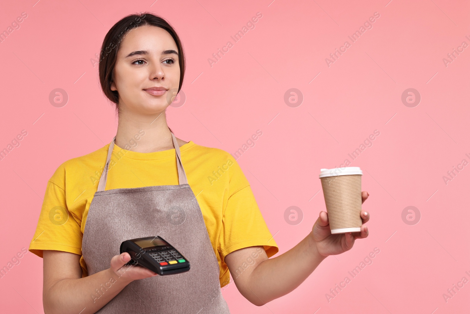 Photo of Girl in apron with paper cup of coffee and payment terminal on pink background. Work for teenagers