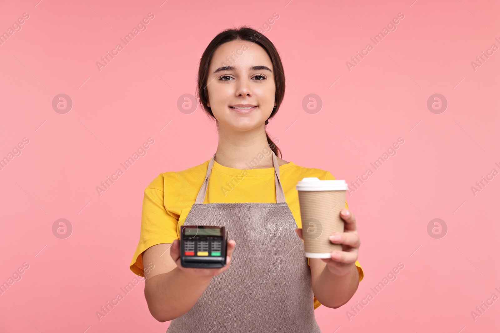 Photo of Girl in apron with paper cup of coffee and payment terminal on pink background. Work for teenagers