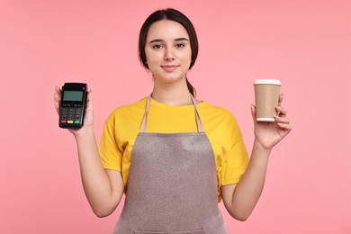 Photo of Girl in apron with paper cup of coffee and payment terminal on pink background. Work for teenagers