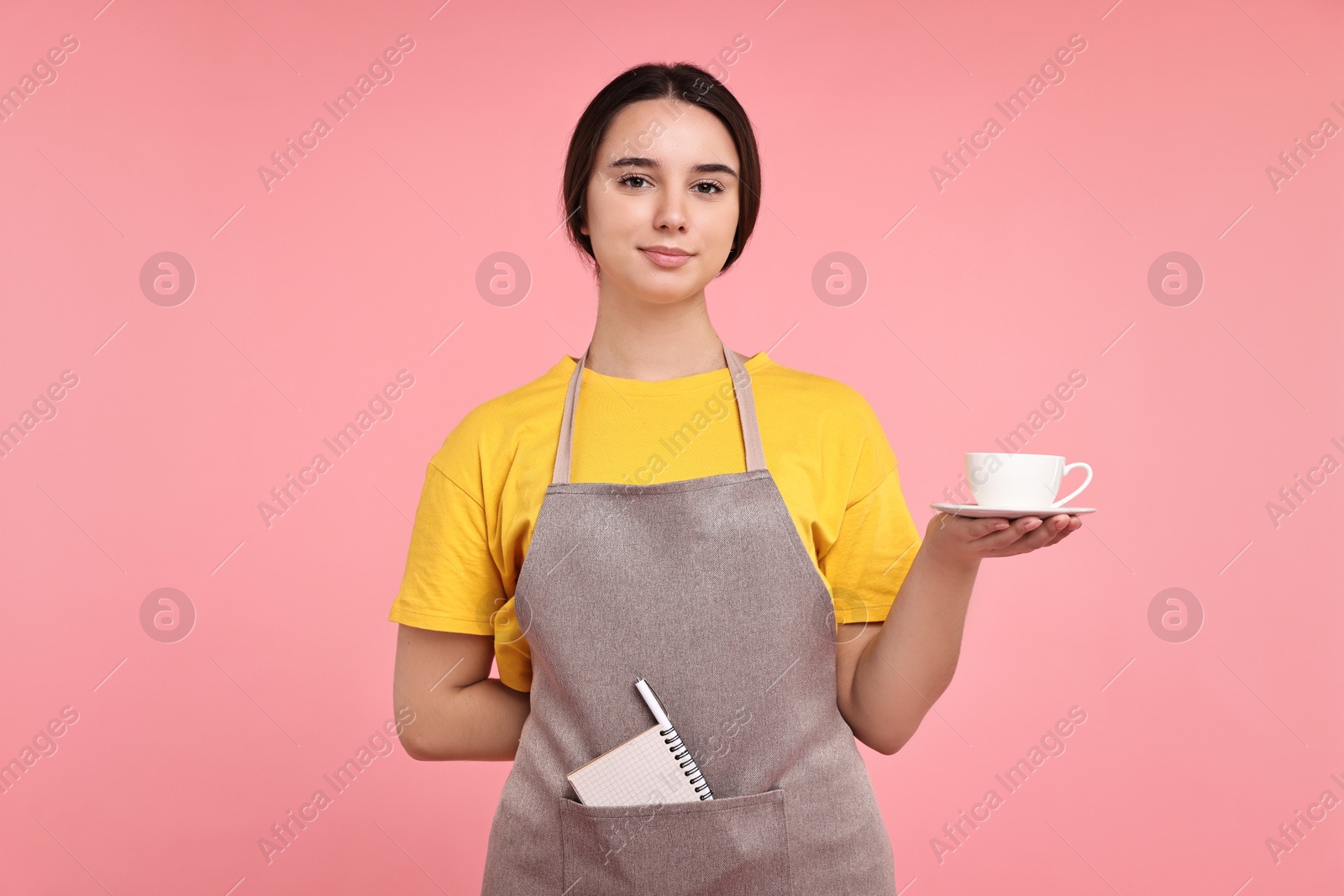 Photo of Girl in apron with cup of coffee on pink background. Work for teenagers