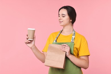 Photo of Girl in apron holding takeaway cup of coffee and paper bag on pink background. Work for teenagers