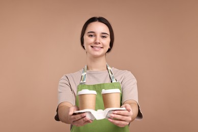 Photo of Girl in apron with takeaway paper cups of coffee on pale brown background. Work for teenagers